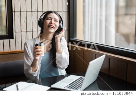 Young professional woman enjoying music while working on a laptop in a modern cafe, promoting productivity and relaxation. 131053944