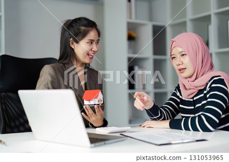 Real estate agent and client discussing property options with a model house in a stylish office environment. Real estate agent and client discussing property options with a model house in a stylish office environment. 131053965