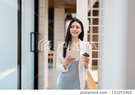 Confident young woman holding a tablet and coffee cup, walking through a stylish office space, embodying modern professionalism. Confident young woman holding a tablet and coffee cup, walking through a stylish office space, embodying modern professionalism. 131053968
