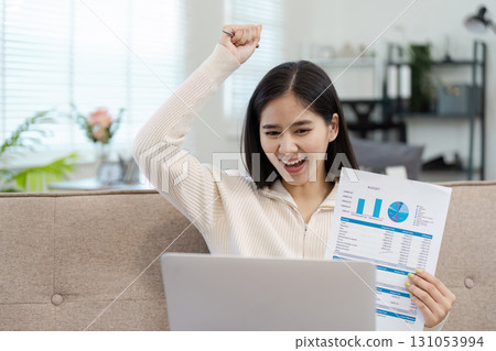 Excited young woman celebrating success while reviewing financial reports at home. 131053994