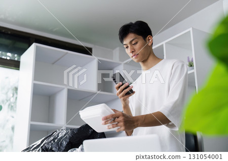 Young man using smartphone while examining recyclable container in eco-friendly workspace, promoting sustainability. 131054001