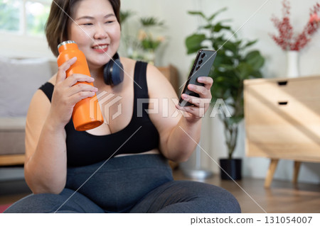 Plus-size woman engaging in wellness, holding a water bottle and checking her phone at home. Plus-size woman engaging in wellness, holding a water bottle and checking her phone at home. 131054007