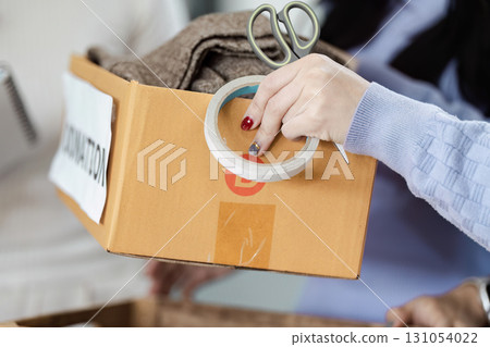 Individual preparing a donation box with clothing items, using scissors and tape for organization and community support. 131054022