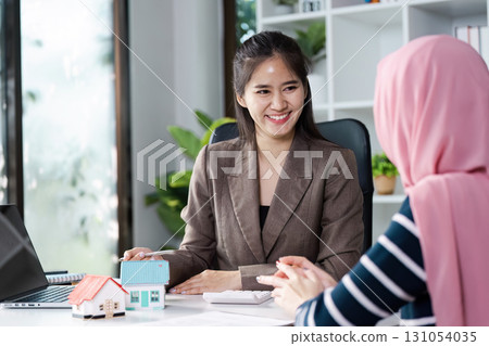 Diverse real estate agent smiling while discussing multiple model houses with client in bright office setting. 131054035