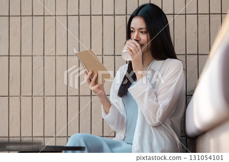 Young Asian woman enjoying coffee while reading a notebook in a cozy cafe setting. Young Asian woman enjoying coffee while reading a notebook in a cozy cafe setting. 131054101