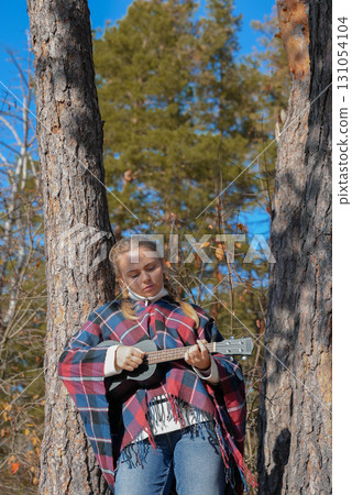 Blonde woman playing guitar ukulele outdoors in autumn forest. Concept of sound therapy, mental health and wellness rituals. Calmness tranquility audio-sensory practices. Aura farming 131054104