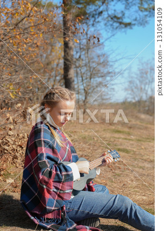 Beautiful blonde girl playing guitar ukulele and singing relaxing enjoying outdoors on sunny autumn day. Hippie woman in boho style training playing musical instrument. Fall leaves Beautiful blonde girl playing guitar ukulele and singing relaxing enjoying outdoors on sunny autumn day. Hippie woman in boho style training playing musical instrument. Fall leaves 131054109