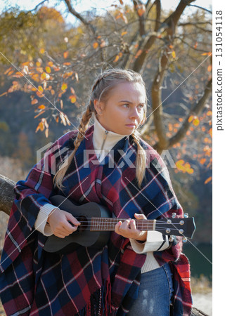 Portrait of smiling caucasian girl plays ukulele in autumnal park. Young woman plays guitar musical instrument outside in nature fall time. Audio music healing Portrait of smiling caucasian girl plays ukulele in autumnal park. Young woman plays guitar musical instrument outside in nature fall time. Audio music healing 131054118