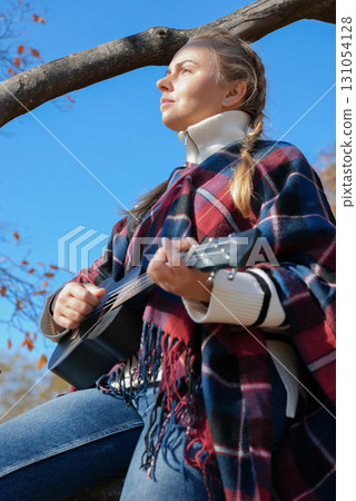 Beautiful blonde girl playing guitar ukulele and singing relaxing enjoying outdoors on sunny autumn day. Hippie woman in boho style training playing musical instrument. Fall leaves Beautiful blonde girl playing guitar ukulele and singing relaxing enjoying outdoors on sunny autumn day. Hippie woman in boho style training playing musical instrument. Fall leaves 131054128