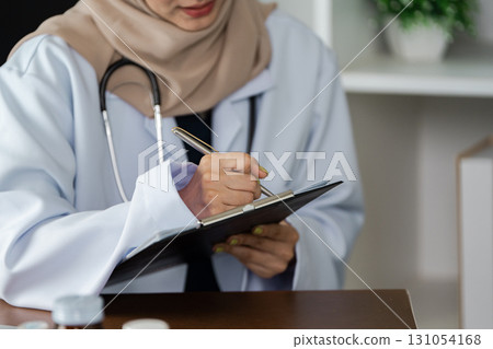 Female doctor in hijab writing notes during a patient consultation, promoting healthcare diversity. 131054168