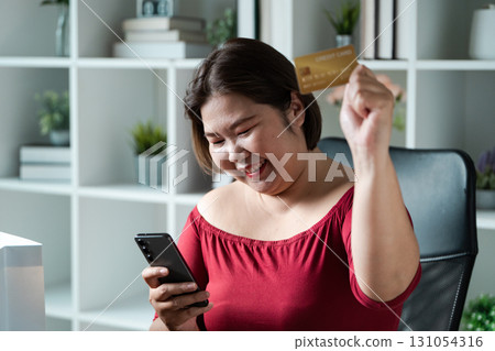Excited woman showing her credit card while shopping online in a cozy workspace. 131054316