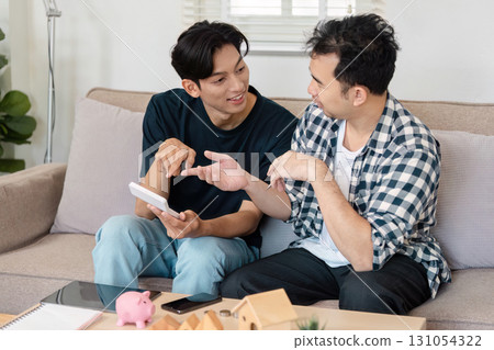 LGBTQ couple enthusiastically discussing savings plans while using a tablet in their cozy living room. Emphasis on financial collaboration and future planning. LGBTQ couple enthusiastically discussing savings plans while using a tablet in their cozy living room. Emphasis on financial collaboration and future planning. 131054322