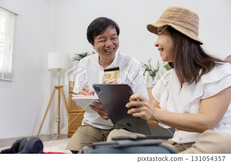 Cheerful Asian couple discussing online shopping, with one holding a credit card and the other using a tablet. 131054357