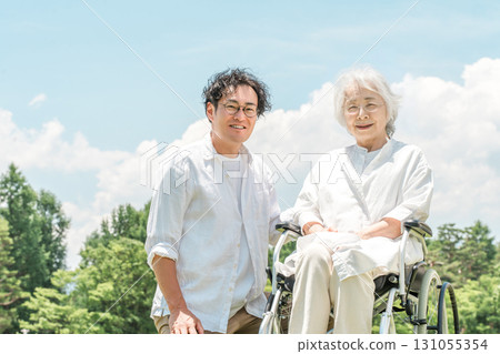 一名坐在輪椅上的老年婦女和一名男子在公園裡協助她（護理、父母和兒童護理、療養院） 131055354