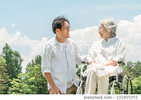 Senior woman in a wheelchair in a park and a man assisting her (caregiving, parent-child, elderly care home) 131055355