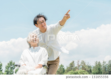 Senior woman in a wheelchair in a park and a man assisting her (caregiving, parent-child, elderly care home) 131055356