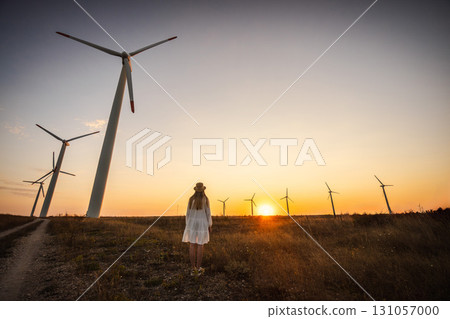 Young woman in white dress standing at sunset near wind turbines, symbolizing renewable energy and harmony with nature. 131057000