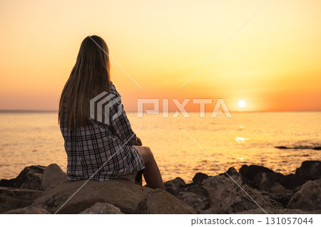 Teenager sitting on rocks at sunset by the sea, enjoying the view 131057044