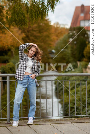 Young woman in trendy autumn outfit posing on a European bridge with golden foliage and a historic cityscape in the background. 131059308
