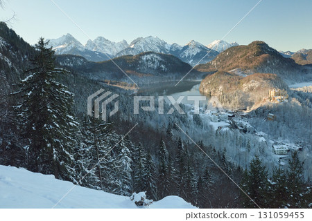 Snowy Mountain Landscape with Iconic Castle Overlooking Serene Lake and Alpine Peaks in Ethereal Winter Wonderland Bathed in Soft Dawn Light 131059455