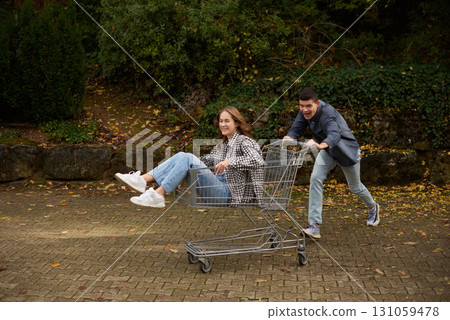 Happy young couple having fun with shopping cart outdoors in autumn park, wearing stylish casual outfits with denim and flannel, laughing and enjoying joyful moment, surrounded by autumn foliage and Happy young couple having fun with shopping cart outdoors in autumn park, wearing stylish casual outfits with denim and flannel, laughing and enjoying joyful moment, surrounded by autumn foliage and 131059478