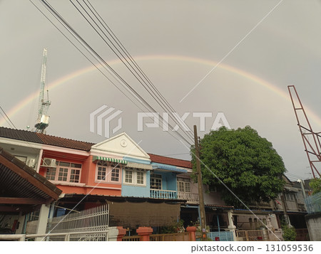 Vibrant rainbow arches across cloudy sky above colorful residential houses with large green tree in foreground, creating peaceful and cheerful atmosphere Vibrant rainbow arches across cloudy sky above colorful residential houses with large green tree in foreground, creating peaceful and cheerful atmosphere 131059536