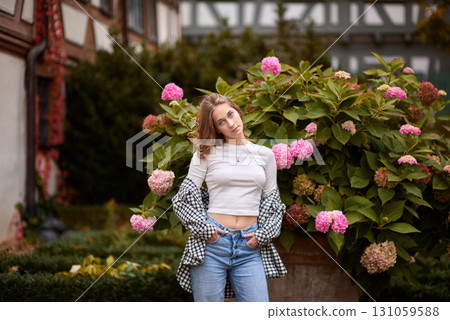 Stylish young woman posing in cozy autumn outfit among blooming hydrangeas in front of traditional half-timbered house in Germany, warm fall colors, outdoor portrait, casual fashion, seasonal Stylish young woman posing in cozy autumn outfit among blooming hydrangeas in front of traditional half-timbered house in Germany, warm fall colors, outdoor portrait, casual fashion, seasonal 131059588