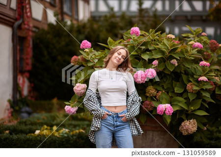 Autumn Fashion Portrait: Stylish Woman Amidst Blooming Hydrangeas at Traditional German Half Timbered House. Cozy Fall Outdoor Scene Showcasing Casual Outfit and Warm Colors in Picturesque Rural 131059589