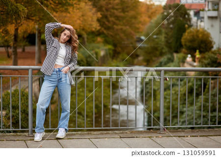 Young woman in trendy autumn outfit posing on a European bridge with golden foliage and a historic cityscape in the background. 131059591