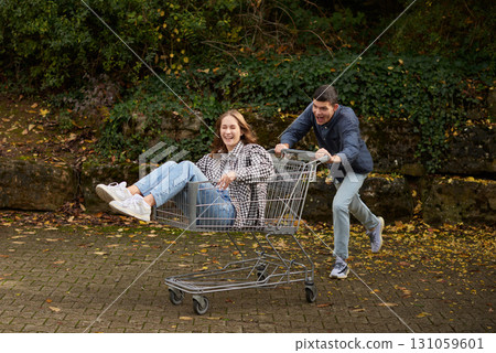 Playful Young Couple's Shopping Cart Adventure in Vibrant Park Setting. Joyful Outdoor Moment Captures Spontaneous Love and Seasonal Casual Style. 131059601
