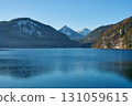Serene Mountainous Lakeside Scene with Calm Reflective Lake, Snow-Dusted Peaks, and Evergreen Trees Under Clear Blue Skies, Featuring a Lone Bird Gliding on Water and a Rocky Cliff with Patches of 131059615