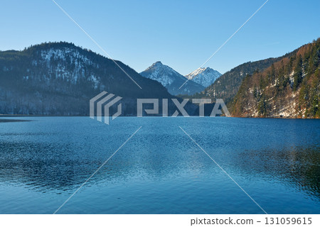 Serene Mountainous Lakeside Scene with Calm Reflective Lake, Snow-Dusted Peaks, and Evergreen Trees Under Clear Blue Skies, Featuring a Lone Bird Gliding on Water and a Rocky Cliff with Patches of Serene Mountainous Lakeside Scene with Calm Reflective Lake, Snow-Dusted Peaks, and Evergreen Trees Under Clear Blue Skies, Featuring a Lone Bird Gliding on Water and a Rocky Cliff with Patches of 131059615