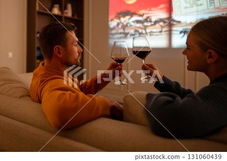 Rear view of couple sharing wine and enjoying film during intimate evening date indoors at home 131060439
