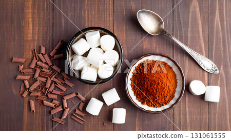 An overhead view of hot chocolate ingredients laid out against a wooden background. In the center are two small bowls: one with cocoa powder, the other with marshmallow cubes. Nearby lies a vintage si An overhead view of hot chocolate ingredients laid out against a wooden background. In the center are two small bowls: one with cocoa powder, the other with marshmallow cubes. Nearby lies a vintage si 131061555