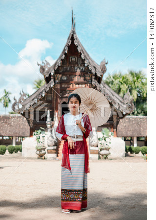 A woman in traditional Thai clothing stands with a parasol in front of an ancient temple, showcasing cultural heritage. 131062312