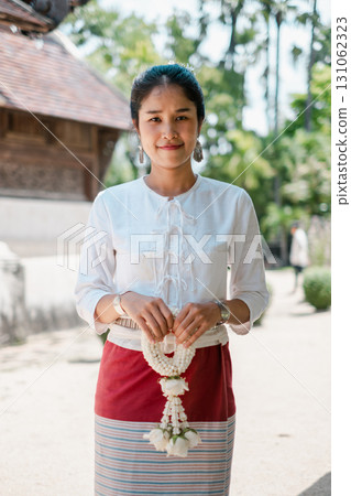 A woman in traditional Thai clothing holds a floral garland, standing outdoors with greenery and traditional architecture in the background. 131062323