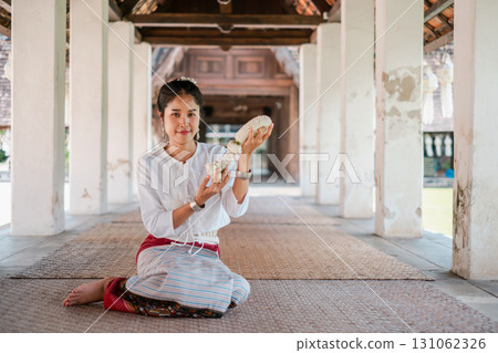 A woman in traditional Thai clothing holds a seashell while sitting in a historic temple corridor, showcasing cultural heritage. 131062326