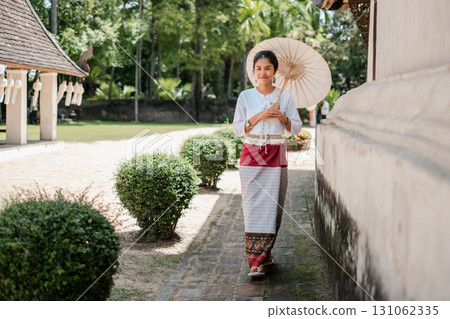 A woman in traditional Thai clothing holding a parasol, walking through a temple courtyard surrounded by greenery. 131062335