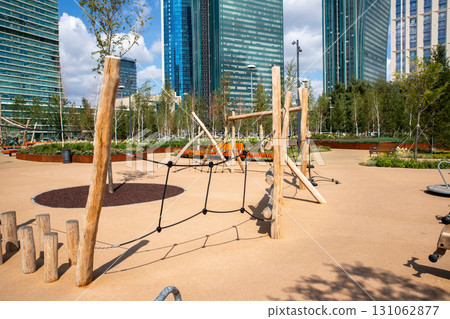 A vibrant playground features wooden structures and swings, set against a backdrop of modern architecture and lush green landscaping during a sunny day. A vibrant playground features wooden structures and swings, set against a backdrop of modern architecture and lush green landscaping during a sunny day. 131062877