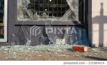 A close-up of a shattered storefront window, with shards of glass lying on the sidewalk. Red bricks used in the destruction lie nearby. The image conveys a sense of destruction, violence, vandalism, a 131064414