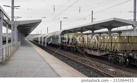 A long freight train with multiple platforms loaded with large containers or crates sits at a railway station under a gray sky. A section of the platform with a canopy is visible in the foreground, cr 131064415