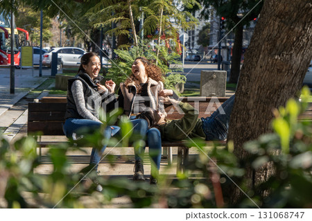 Three young women laughing together on bench in city park Three young women laughing together on bench in city park 131068747
