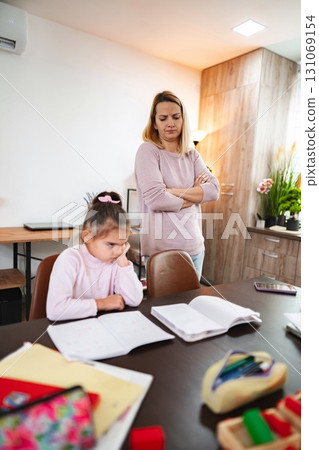 Mother watches her daughter focus intently on homework at home 131069154