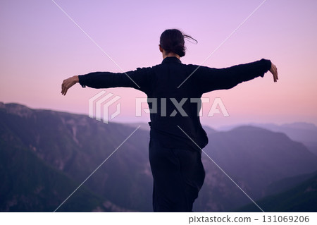 Woman practicing Tai Chi in traditional black outfit on mountain at sunrise, arms outstretched in meditation pose focusing on balance, energy, and mindfulness in nature. 131069206