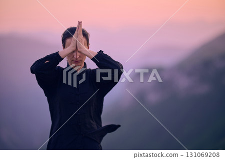 Woman practicing Tai Chi in traditional black outfit on mountain at sunrise, arms outstretched in meditation pose focusing on balance, energy, and mindfulness in nature. 131069208