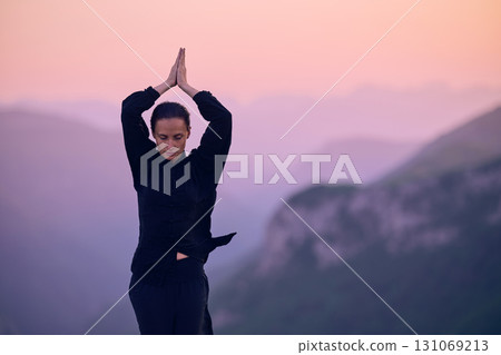 Woman practicing Tai Chi in traditional black outfit on mountain at sunrise, arms outstretched in meditation pose focusing on balance, energy, and mindfulness in nature. 131069213