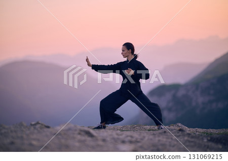 Woman practicing Tai Chi in traditional black outfit on mountain at sunrise, arms outstretched in meditation pose focusing on balance, energy, and mindfulness in nature. 131069215