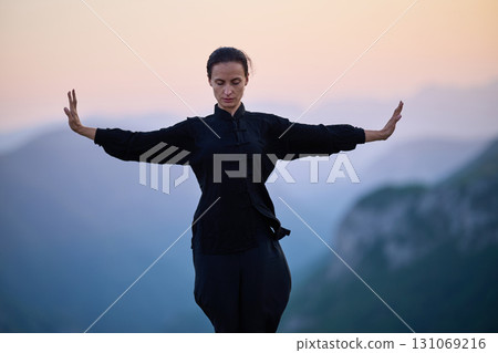 Woman practicing Tai Chi in traditional black outfit on mountain at sunrise, arms outstretched in meditation pose focusing on balance, energy, and mindfulness in nature. 131069216