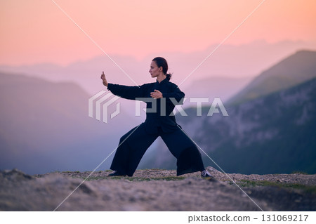 Woman practicing Tai Chi in traditional black outfit on mountain at sunrise, arms outstretched in meditation pose focusing on balance, energy, and mindfulness in nature. Woman practicing Tai Chi in traditional black outfit on mountain at sunrise, arms outstretched in meditation pose focusing on balance, energy, and mindfulness in nature. 131069217
