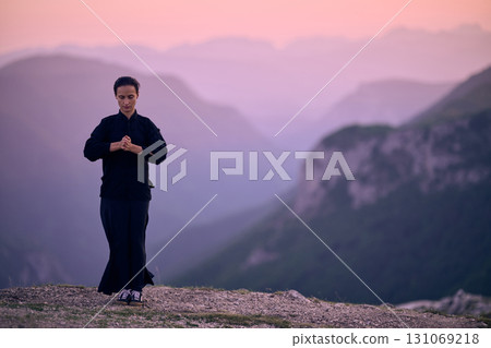 Woman practicing Tai Chi in traditional black outfit on mountain at sunrise, arms outstretched in meditation pose focusing on balance, energy, and mindfulness in nature. 131069218